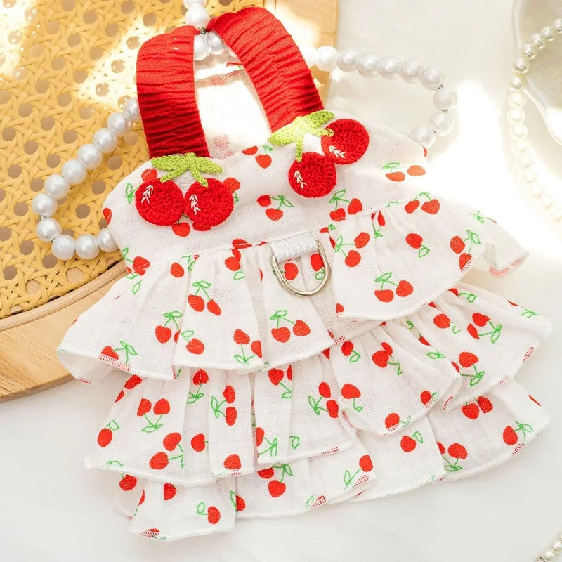 White cherry-print pet dress with red ruffled straps and embroidered cherries by PetoPop, featuring tiered ruffles and a silver D-ring closure, displayed flat with pearls and woven basket.
