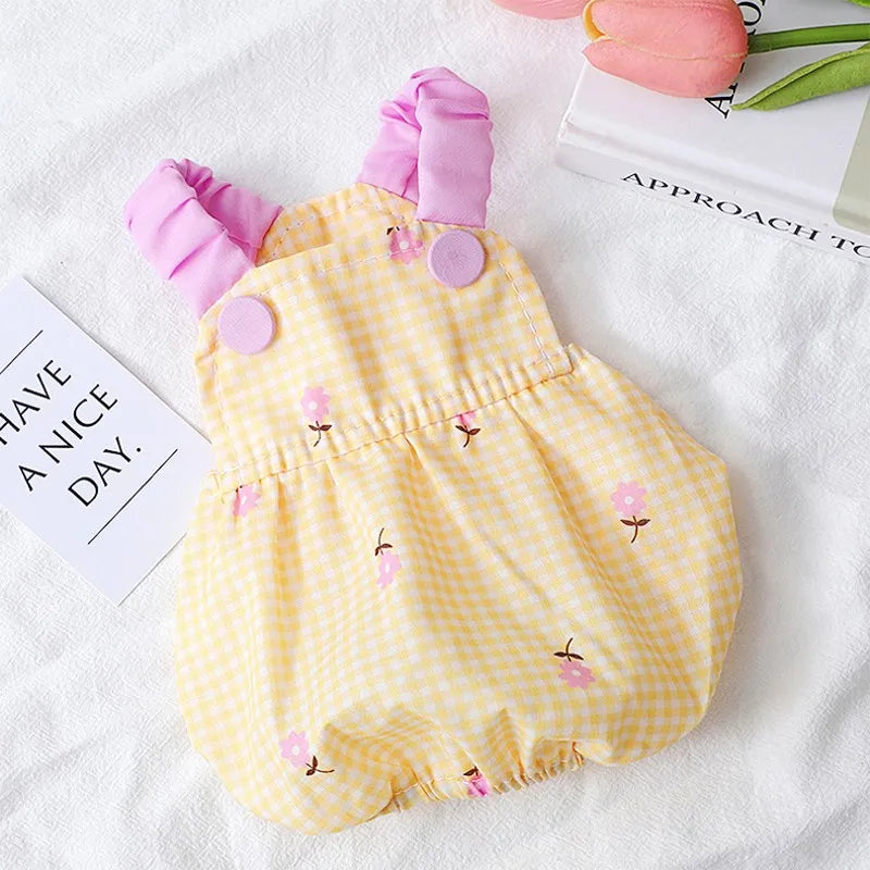 Yellow gingham dog and cat dress with pink floral embroidery and ruffled straps by PetoPop, laid flat on white fabric with decorative props.